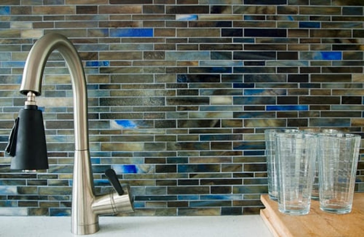 Glass mosaic kitchen backsplash in blue and bronze tones behind a brushed nickel faucet